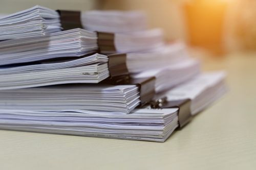 Education concept: Close and selective focus of unfinished homework assignment stack archives with binder paper clips on teacher’s desk in university waiting to be evaluated and scored. Stack of white paperwork achieves with black binder paper clips.